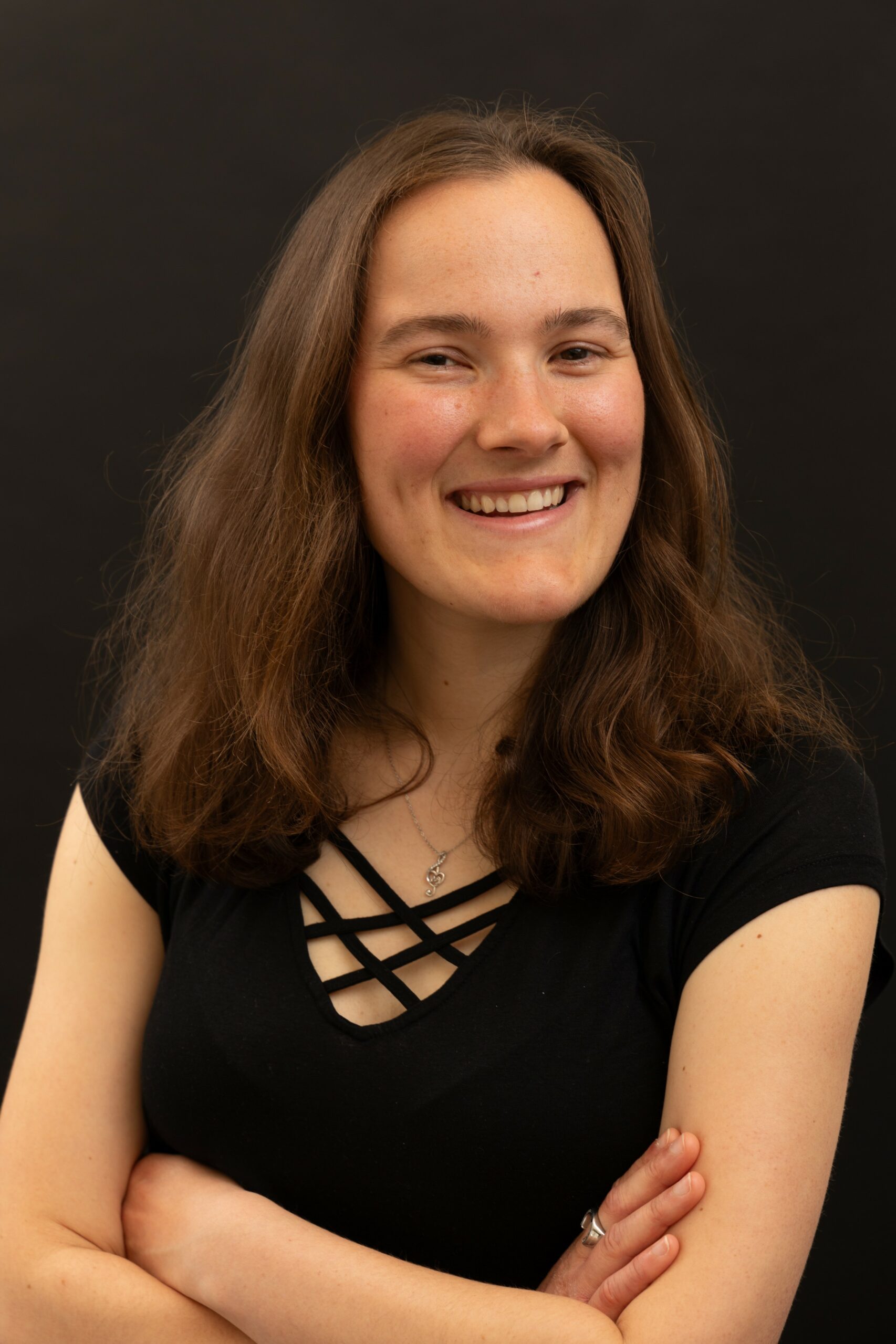 Photo portrait of a woman. She is smiling. She has light brown hair and she is wearing a black t-shirt. She is crossing her arms. She looks professional.