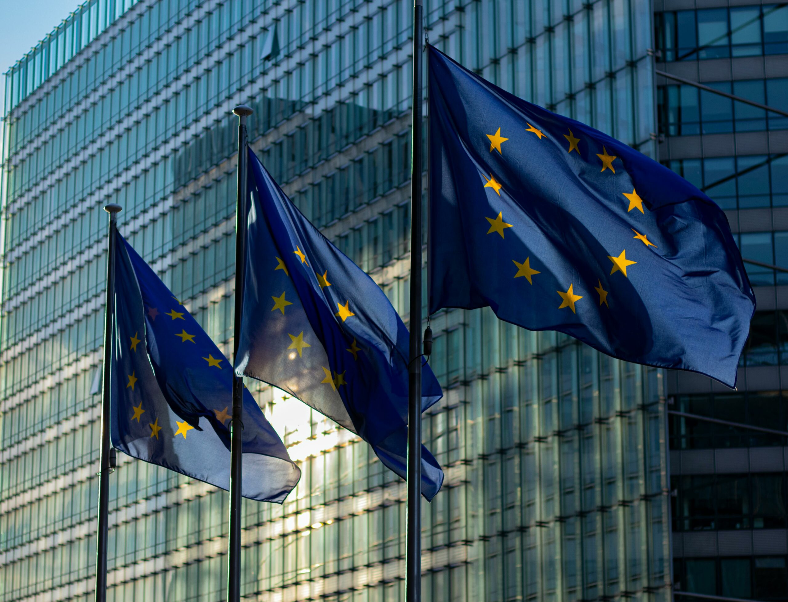 Three European Union flags waving in the wind, set against a backdrop of a modern glass building, possibly the builiding of the European Commission.